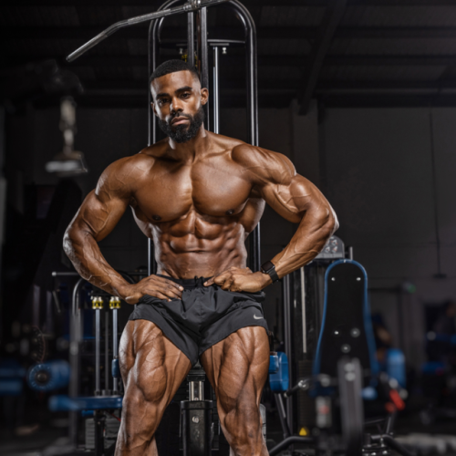 Muscular bodybuilder posing in a gym with defined abs and quads, hands on hips.