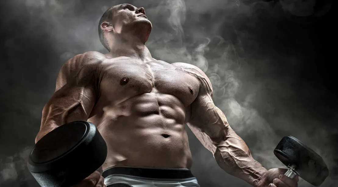 Strong male athlete holding dumbbells against a smoky background, upper body flexed.