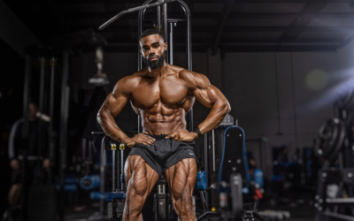 Muscular bodybuilder posing in a gym with defined abs and quads, hands on hips.