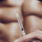 A detailed close-up of a syringe used for performance enhancement with a blurred view of defined abdominal muscles in the background.