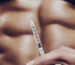 A detailed close-up of a syringe used for performance enhancement with a blurred view of defined abdominal muscles in the background.